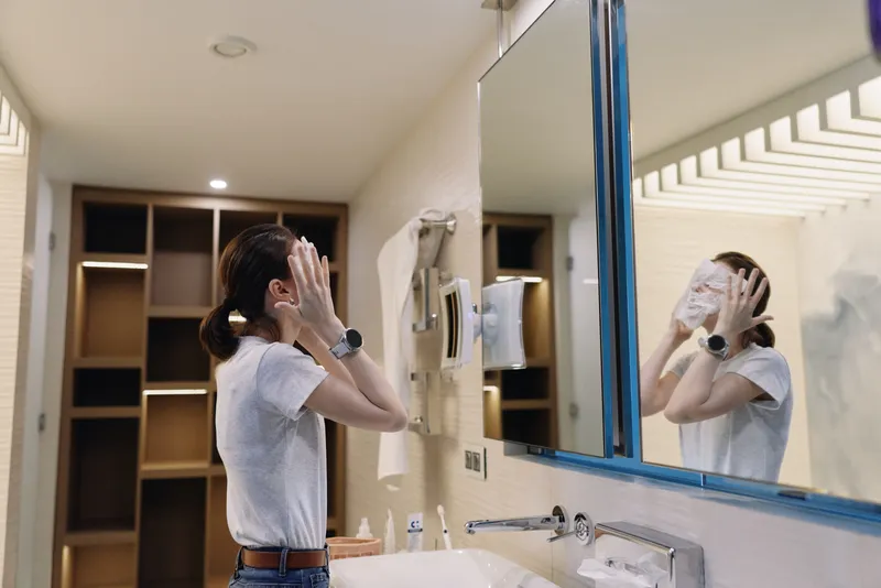Skincare routine in bathroom with a woman facing a mirror, applying product on her face, casual attire, clean sink area, soft lighting, reflective glass, a calm self care moment