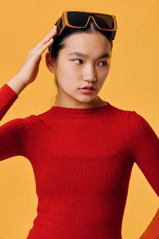 Young woman in red sweater adjusts sunglasses on yellow background, looking sideways with thoughtful expression. Studio portrait with bright colors and casual fashion style.