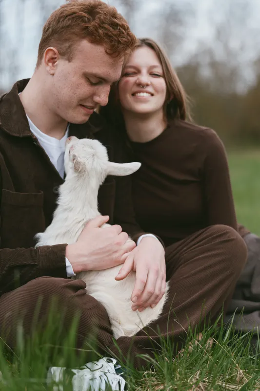 A young man gently holds a white baby goat while sitting on grass with a smiling young woman nearby in casual brown clothing during a relaxed outdoor lifestyle moment.