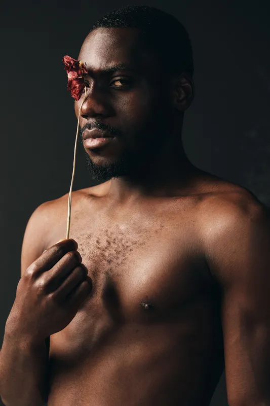 Creative conceptual portrait of African man with bare chest holding dried flower in front of face, artistic visual style with dark background and moody lighting.