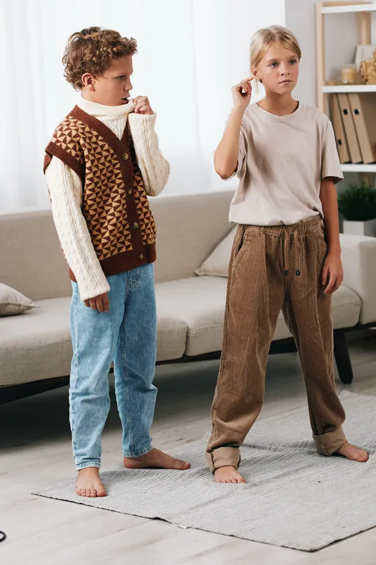 Two young children stand indoors on a light rug, wearing cozy knitwear and casual pants, capturing a quiet family moment of comfort, curiosity, and gentle interaction.