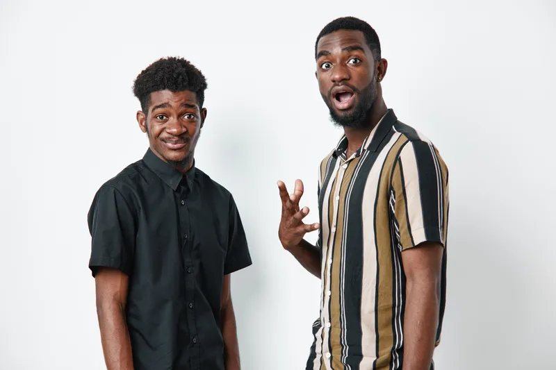 Two young black men with dark skin posing on white background one in black shirt and the other in striped shirt showing surprised and skeptical expressions for casual portrait.