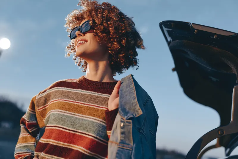 Woman lifestyle outdoors, curly hair and sunglasses, denim overalls and striped sweater, standing beside a piano, bright sunny sky, relaxed smile, music focused moment