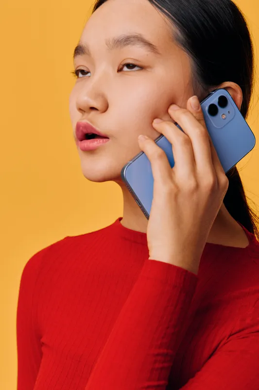 Young woman in red sweater holding smartphone to ear with thoughtful expression on orange background. Closeup portrait of female using mobile device for communication and conversation.