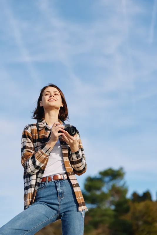 woman photographer outdoors camera blue sky casual, confident stance as she captures moments in daylight, wearing plaid shirt and denim while looking up