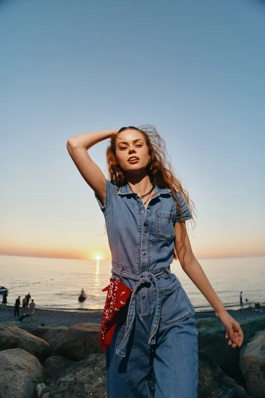 young woman in denim jumpsuit stands on rocky beach at sunset with relaxed expression and casual pose, calm ocean and pastel sky in background