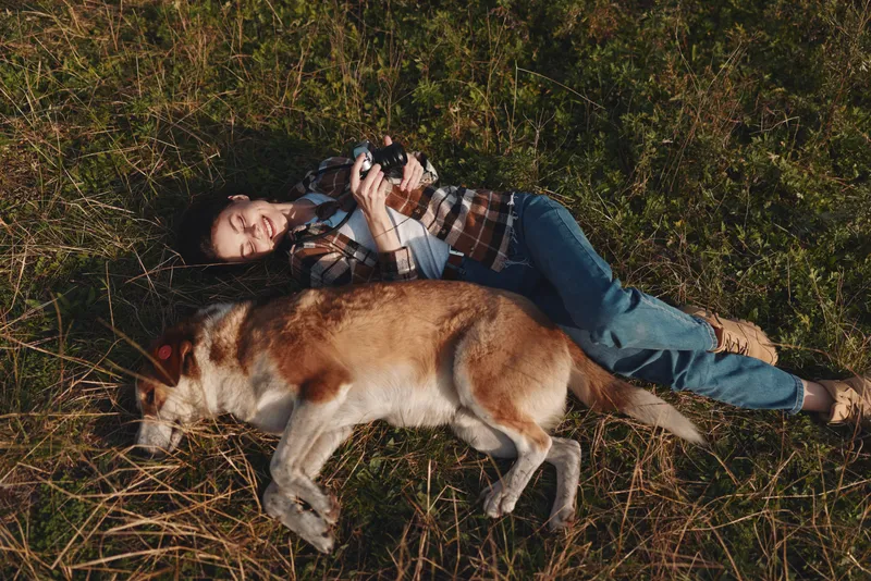 Person lying on grass outdoors with a dog, relaxed and in a peaceful moment, enjoying nature and sunlight amidst tall vegetation, casual clothing and a calm companionship