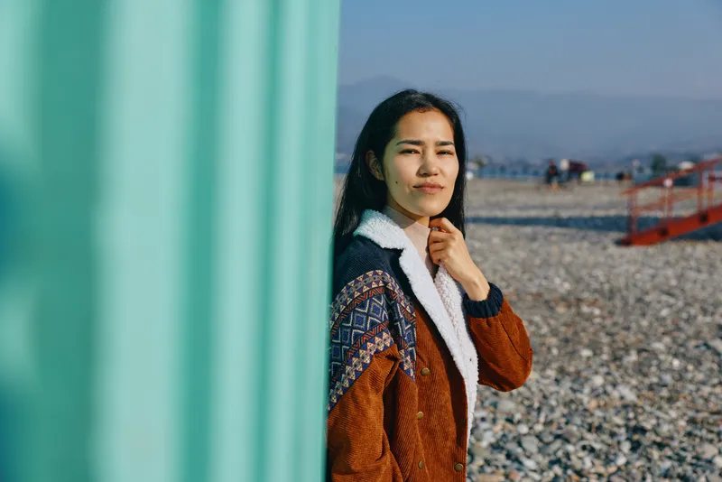 Woman outdoor wall sea coastline daylight cardigan scarf standing beside a turquoise wall by a pebbled waterfront, gazing into distance under bright daylight