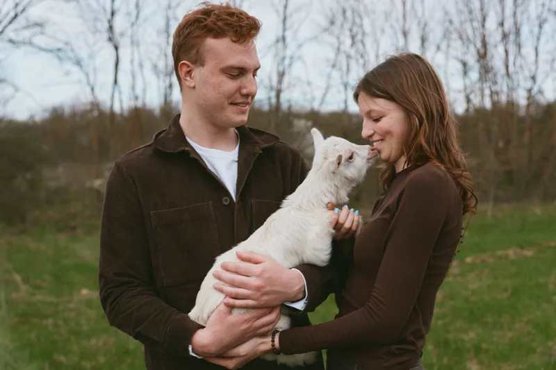 Young couple enjoys a tender moment outdoors as the man holds a white baby goat while the woman smiles and interacts with the animal in a natural lifestyle setting.