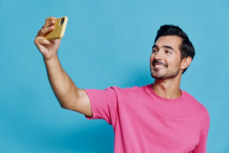 Smiling man in pink t-shirt takes selfie with mobile phone smiling happily at camera against bright blue background. Modern lifestyle concept.