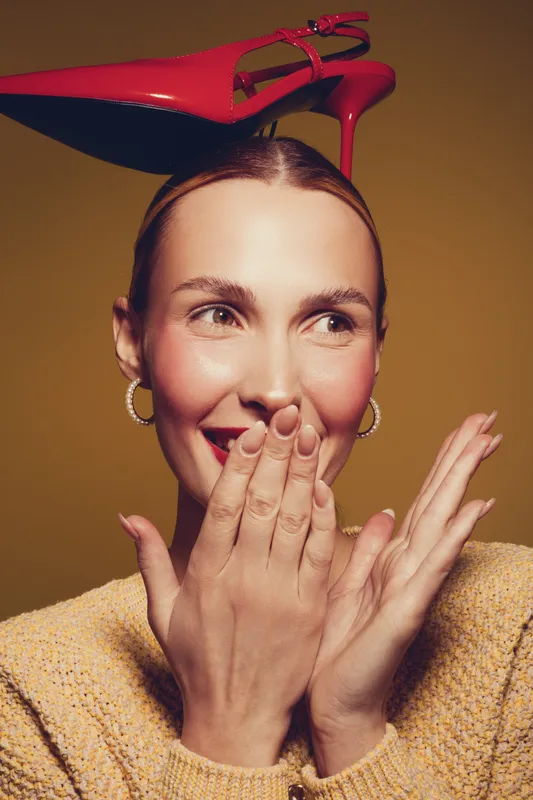 Smiling woman with red high heel shoe on head covers mouth with hands. Studio portrait on brown background. Concept of fashion, fun, and playful mood with creative styling and accessories.