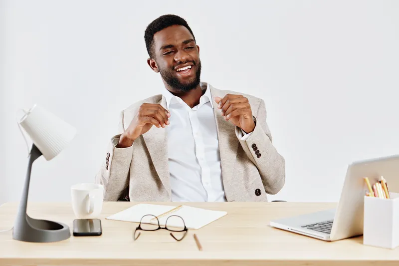 Confident young African American man in beige blazer and white shirt smiling happily while sitting at desk in modern office, holding jacket straps, with glasses, laptop, and office supplies, concept o