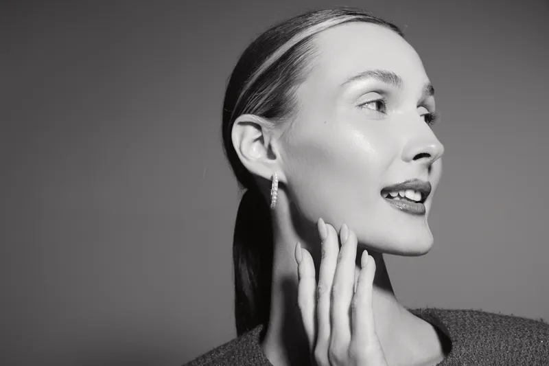 Profile of a young white woman with fair skin touching her cheek, smiling softly against a plain background.