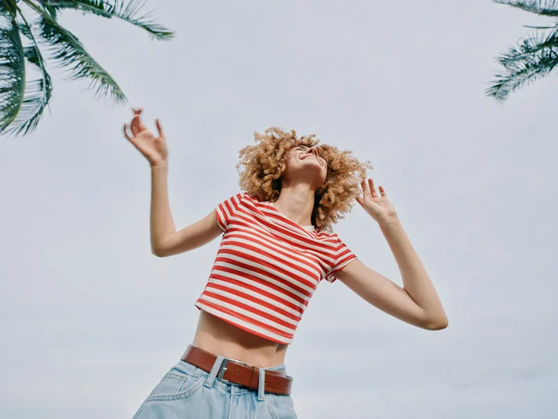 Joyful dancer leaps beneath palm trees, wearing a red and white striped crop top and denim shorts, hair bouncing as sunlight highlights a carefree summer moment.