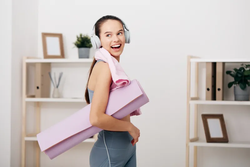Woman fitness enthusiast wearing headphones, carrying a pink yoga mat and towel, smiling in a bright home studio with shelves, plants and framed art.