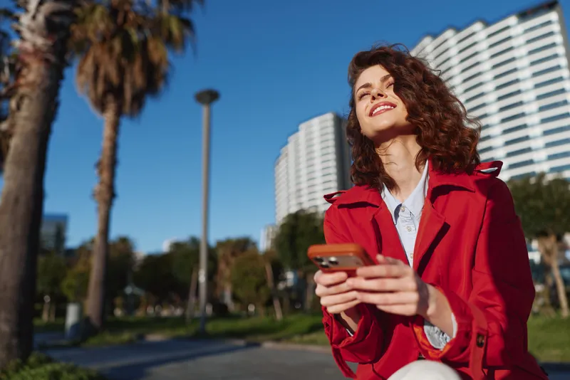 Young woman with curly hair wearing a red coat smiles happily while holding a smartphone outdoors on a sunny day near modern buildings and palm trees.
