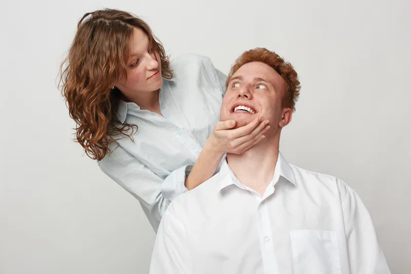 Young woman gently holds smiling man’s face while looking at him. Both wear white shirts against plain background. Concept of affection, care, and close relationship.