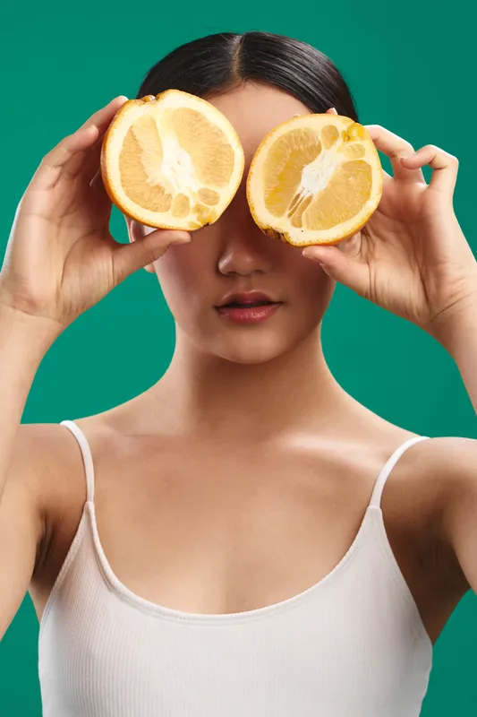 asian woman playfully holds lemon halves over her eyes in a bright studio setting, creating a whimsical portrait that emphasizes freshness, curiosity, and a vibrant mood.