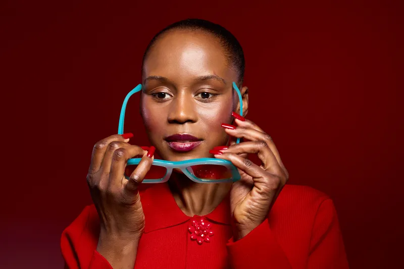 Portrait of a confident black woman with dark skin holding blue glasses in front of her face wearing a red sweater. Studio shot on red background with vibrant fashion concept.