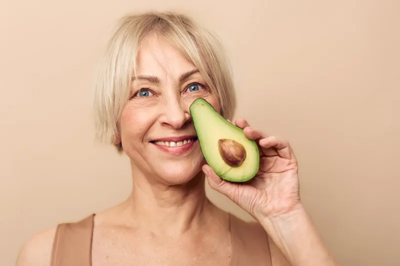 Smiling mature woman holds half of fresh avocado near face on beige background. Healthy lifestyle concept with natural organic fruit for nutrition and skincare at home or studio.