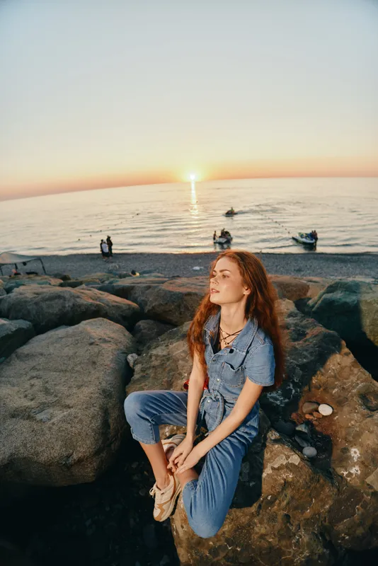 lifestyle woman in modern boho-western denim street style sitting on rocks at sunset with warm film color tones and relaxed mood near ocean shore