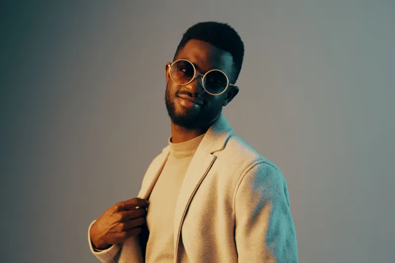 Stylish African man in round sunglasses and a light blazer, smiling confidently against a gradient background, showcasing modern fashion trends and vibrant style