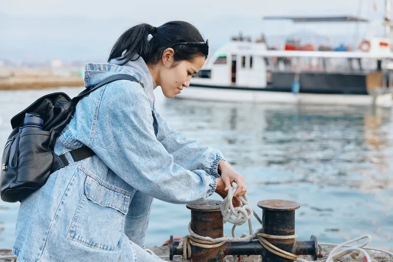 Woman by a harbor mooring rope, denim jacket, preparing knot near water, boat in background, travel scene, casual outdoor activity, salty air, calm maritime atmosphere, glow of sunset and distant