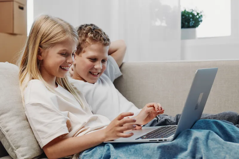 Two happy children sitting on a couch using laptop together in bright living room. Concept of friendship, technology, and leisure at home.