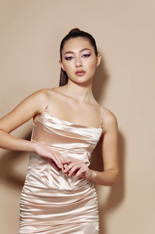 Young woman in a satin slip dress poses with hands clasped softly, wearing bold creative pink eye makeup. Neutral background highlights fashion and beauty style.