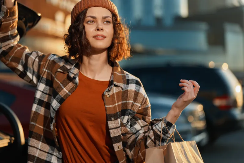 woman shopping bags urban smile candid golden authenticity: young woman in plaid jacket and beanie carrying paper bags on city street, candid lifestyle with golden hour glow, mindful living and