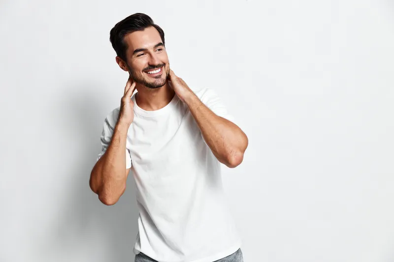 Smiling handsome young man wearing a casual white t-shirt, looking at the camera, with a joyful expression and a charming pose against a plain light background.