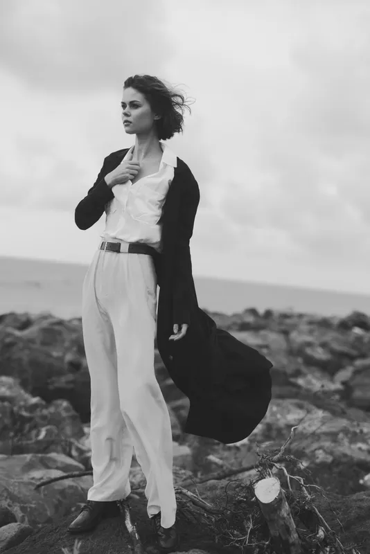 Fashion model stands on a rocky coast in a flowing coat and light trousers, wind catching her hair and coat, black and white photography capturing moody coastal drama