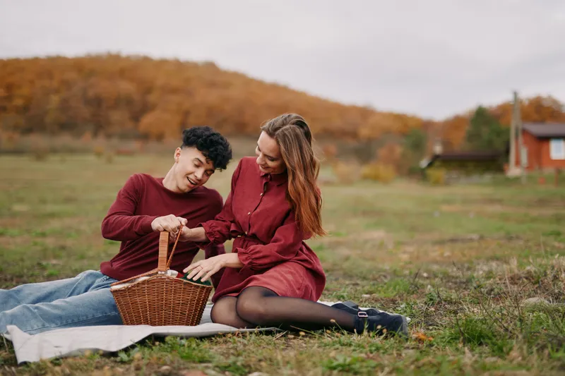 A candid outdoor picnic scene of a young couple sharing a wicker basket on a blanket in an autumn field, conveying authenticity and warm genuine connection through natural poses.