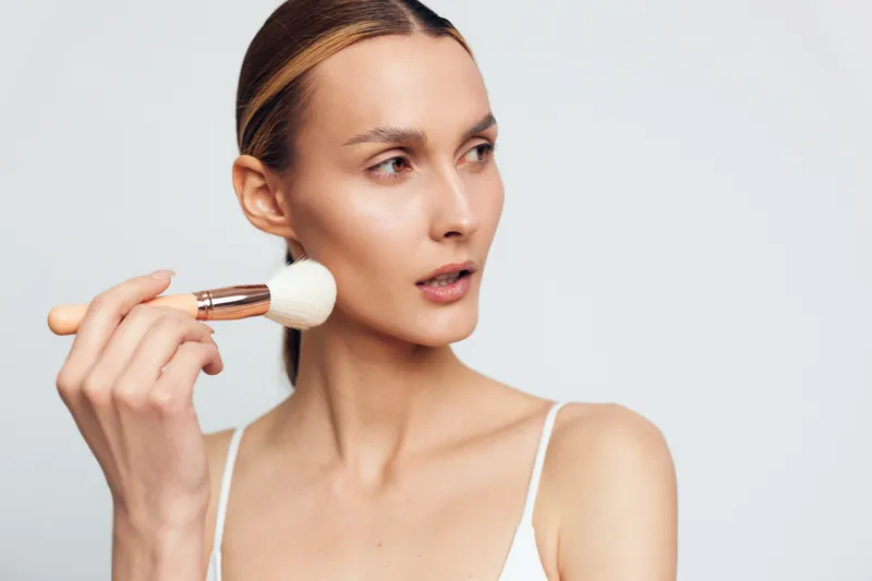 Woman applies makeup with a brush for skincare beauty care, focused face on clean white background, close up grooming routine, natural calm self confidence today