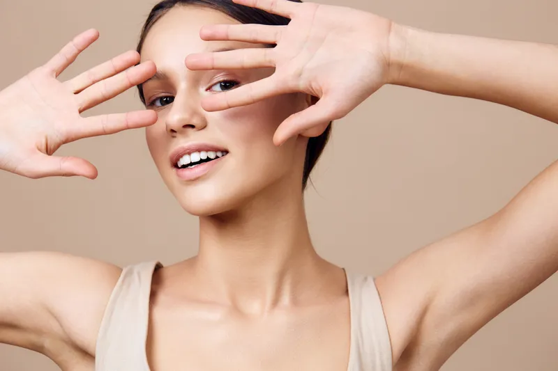Young smiling woman playfully covering her face with hands, showcasing natural beauty against a beige background, emphasizing a healthy lifestyle and positive emotions