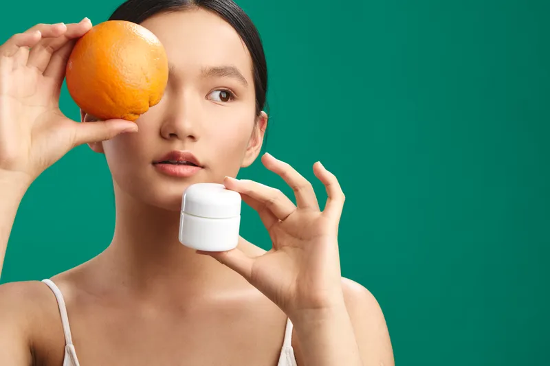 asian woman holds orange near eye in a studio portrait, exuding curiosity and calm while presenting a fresh wellness and beauty vibe against a clean neutral background.