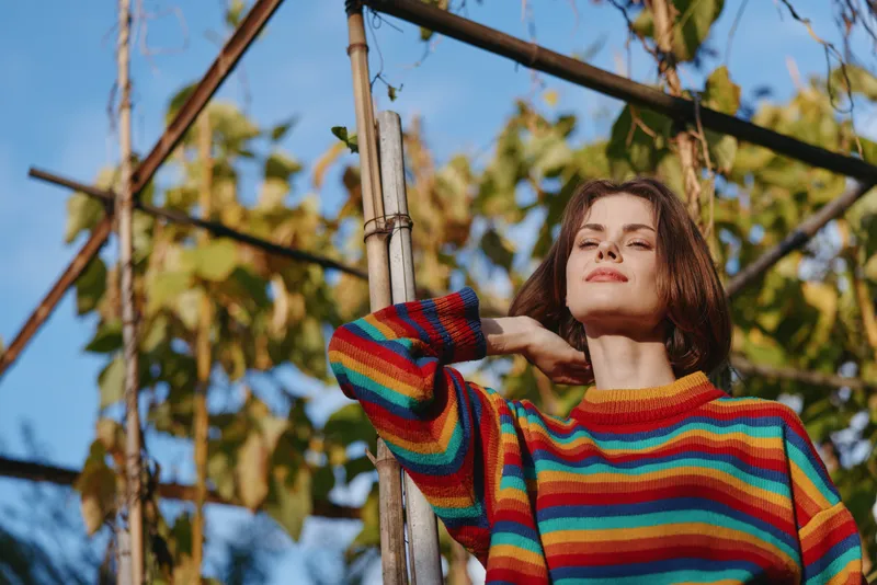 Woman in a colorful striped sweater leaning against a wooden trellis in an outdoor garden, brown hair, relaxed expression and sunlight on her face, lifestyle portrait and casual fashion.
