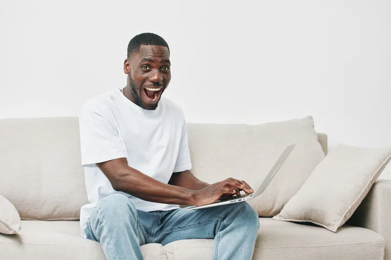 Excited young Black man enjoying work on a laptop at home, wearing casual white t shirt and jeans, expressing happiness and engagement in a modern, light filled environment