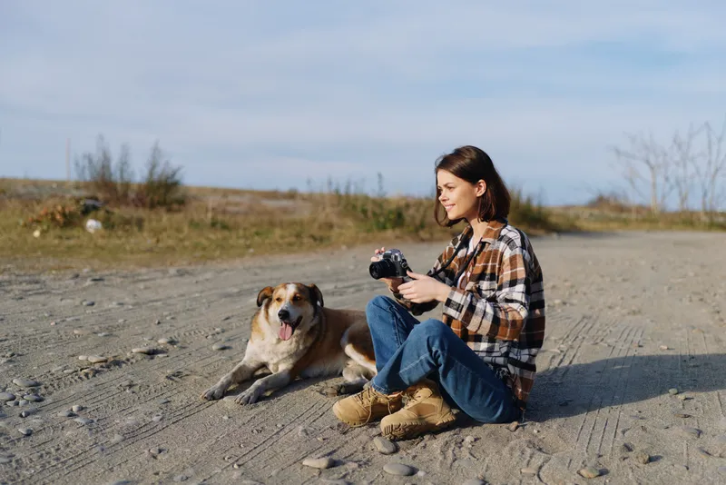 A woman sits on a sandy trail with a camera, a dog resting nearby, enjoying a relaxed outdoor photography moment under a clear sky and wide open scenery