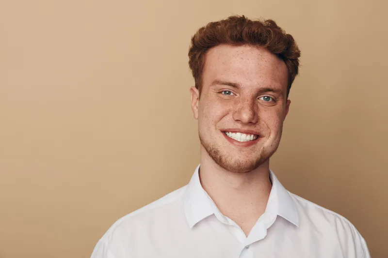 Smiling young man with curly hair and freckles wears white shirt against beige background. Portrait of confident male model with natural expression and casual style in studio lighting.