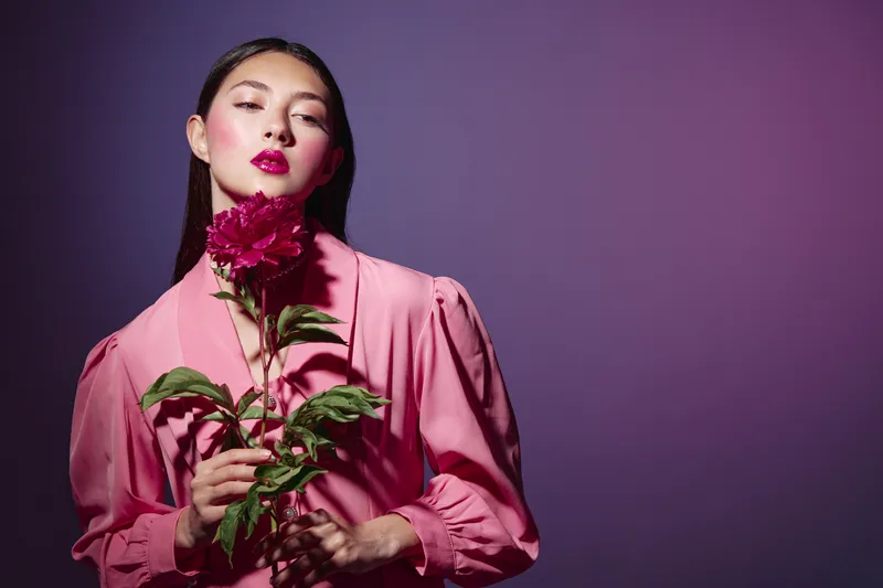 Young woman with striking makeup holds a large dark pink flower, wearing a stylish pink blouse on purple background. Creative beauty fashion portrait concept.
