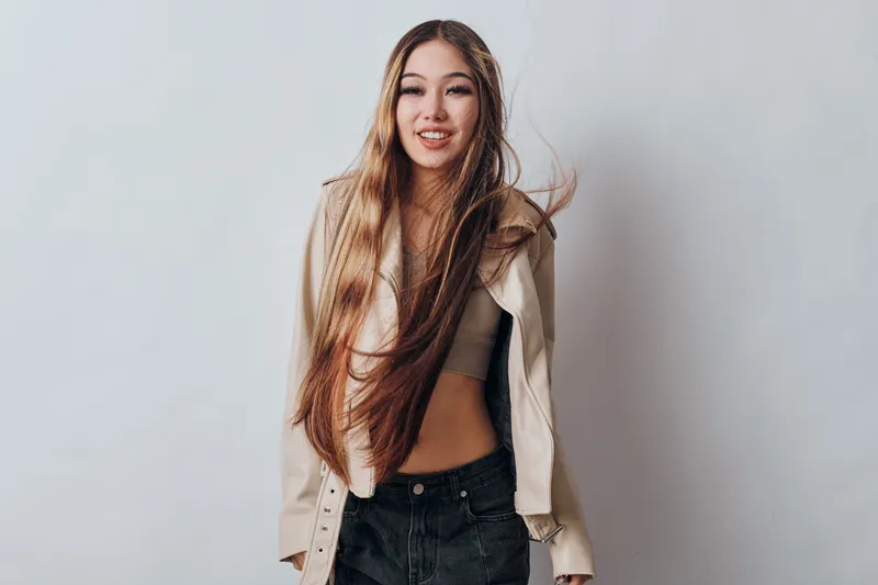Young woman with long hair wearing casual jacket and black pants stands smiling against gray background in studio, showing confidence and relaxed fashion style.