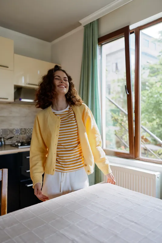 A smiling young woman in a yellow cardigan stands in a bright kitchen by a window, wearing a striped shirt and white pants, enjoying a relaxed morning at home, sunlit and cozy.