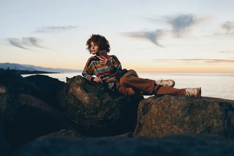 Woman lifestyle portrait sitting on rocks by the sea at sunset, outdoor casual outfit in warm golden light. Relaxing pose, curly hair, ocean horizon.