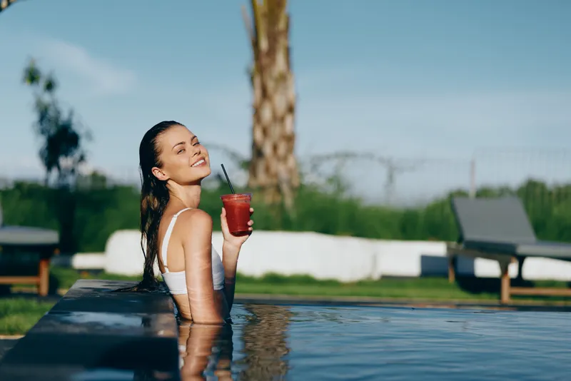 Lifestyle portrait of a smiling woman in a pool, holding a drink with a straw, enjoying summer relaxation outdoors in swimwear under sunshine, vibrant mood by the water.