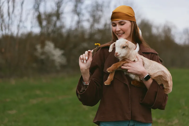 A joyful woman in a brown jacket holds a goat kid outdoors, presenting a small flower. A candid lifestyle moment highlighting rural bonding, animal companionship, and playful connection in a natural