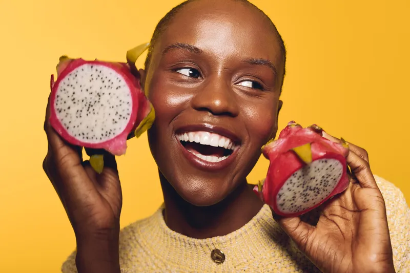 Happy African woman with dark skin holding two halves of dragon fruit near her face smiling on yellow background. Concept for healthy lifestyle, fresh fruit, and natural beauty.