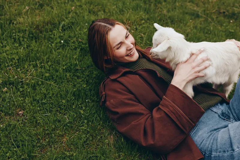 A woman lies on green grass, smiling, cradling a small goat kid in her arms. An outdoor lifestyle moment showcasing warmth, trust, and connection between person and animal in a calm scene.