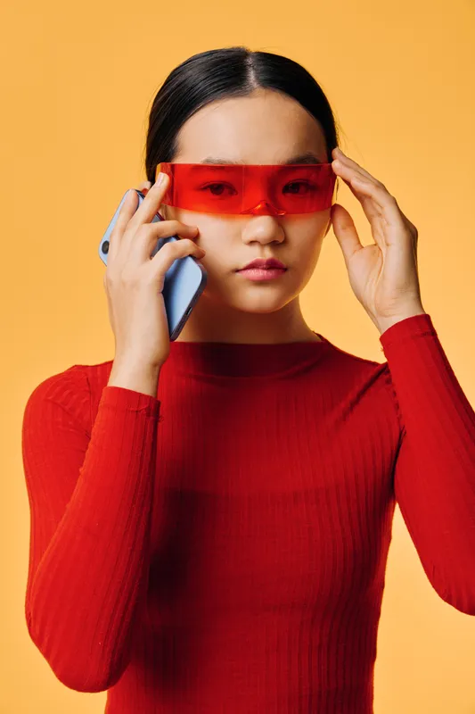 Young woman in red sweater and red futuristic glasses holds smartphone to ear on orange background. Stylish female with modern accessories posing for portrait with minimal expression.