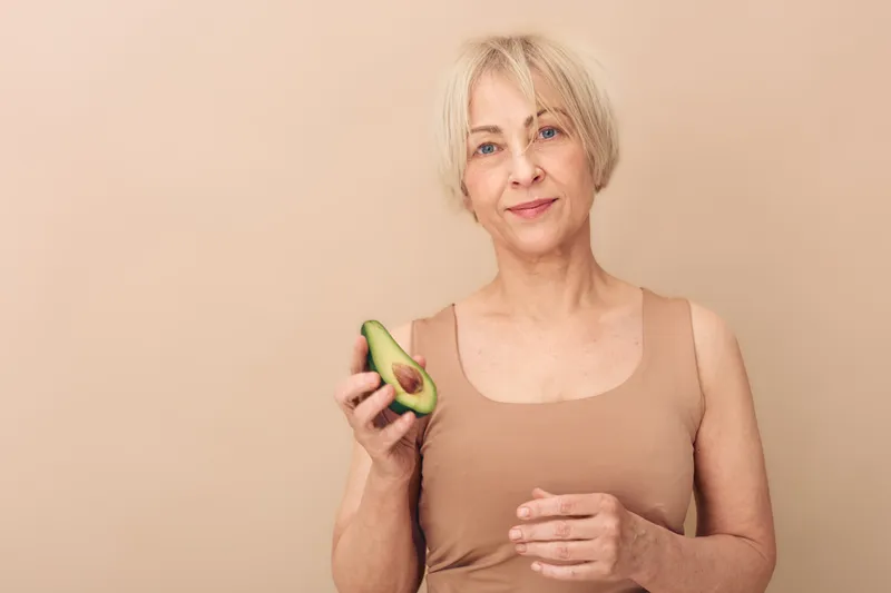 Mature woman with short blonde hair holds half an avocado, smiling gently. Beige background and neutral clothing emphasize natural wellness and healthy lifestyle concepts.